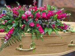 A splendid and rich combination of magenta coloured garden flowers arranged beautifully on an oak coffin with gold handles.   