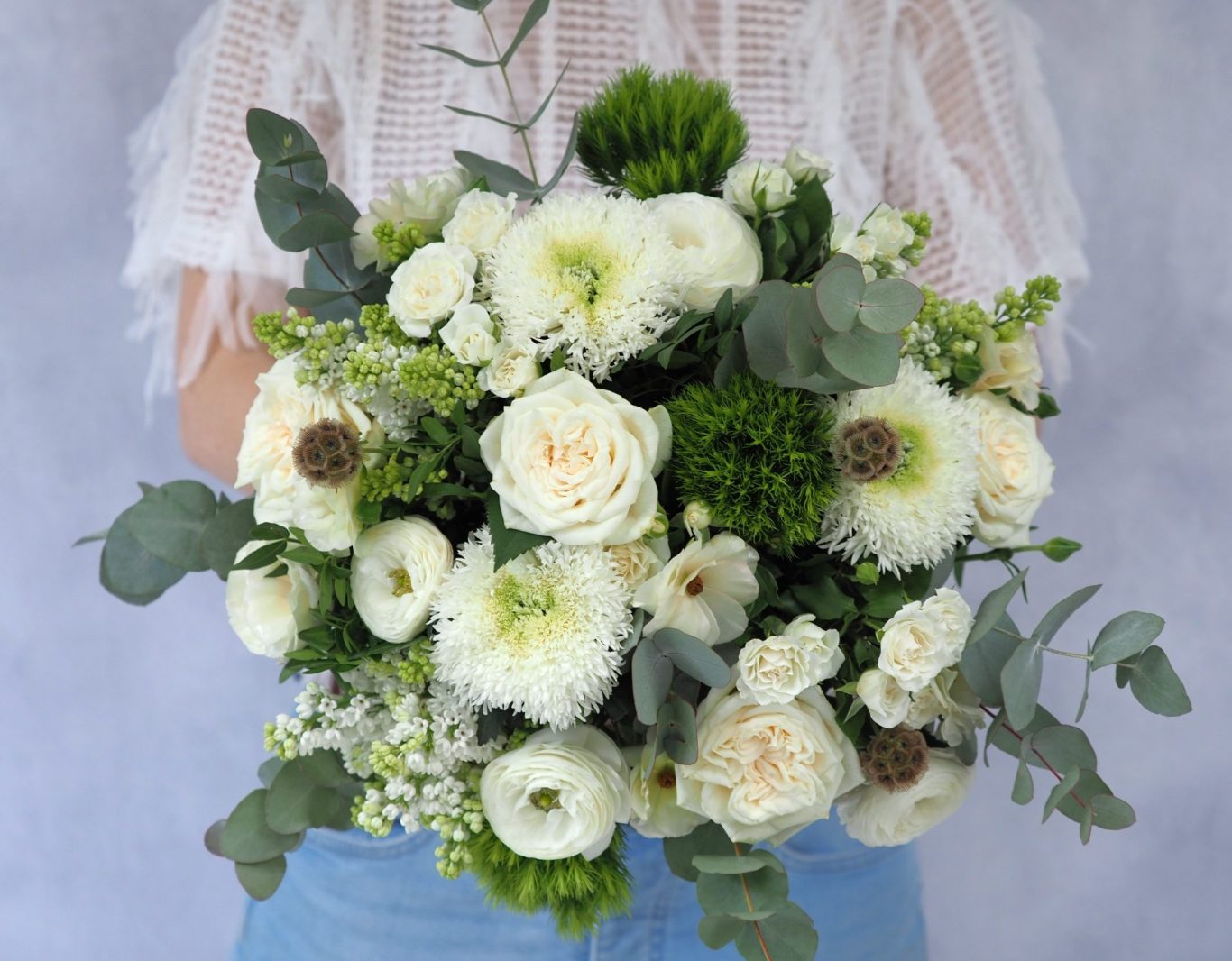 A selection of white, cream and green flowers including white lilac, scented cream garden roses and eucalyptus. Hand tied into a structured and abundant styled bouquet.   