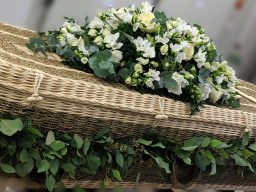 A wicker coffin decorated with a garland made from eucalyptus and textured greenery. Adorned with a double ended spray of elegant scented white flowers.