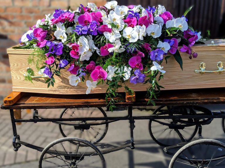 A cascading double ended funeral flowers spray of phalaenopsis orchids in rich pinks purples and whites, arranged elegantly on a beautiful oak coffin with gold handles. 